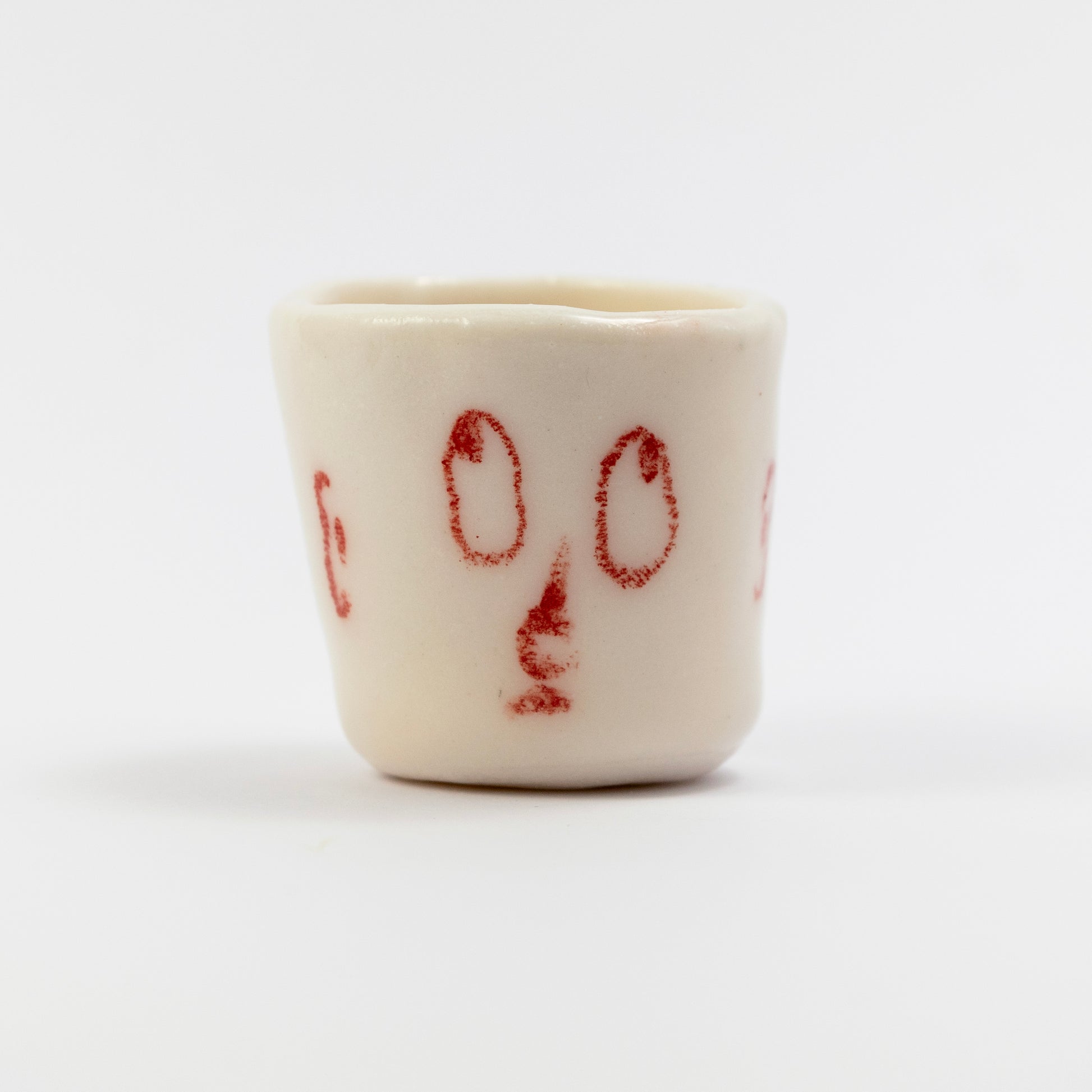 Small white porcelain cup with red underglaze pencil drawing of two oval eyes and a simple nose shape.