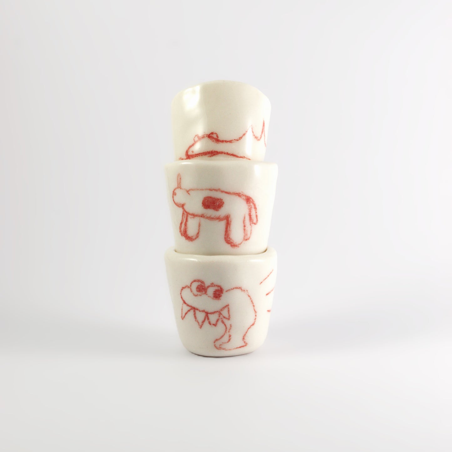 3 ceramic shot glasses stacked on top of each other. There is red hand-drawn doodles on each glass. Bottom: Monster with large spikey teeth. Middle: Horse like creature with circle on it's stomach. Top: A sleeping dinosaur.