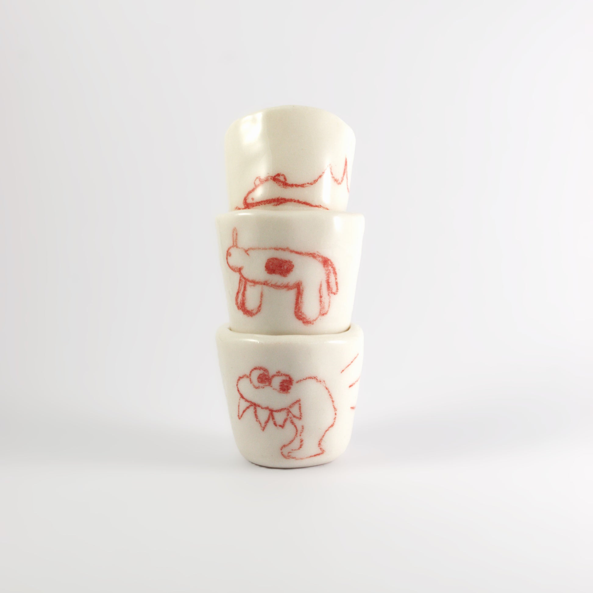 3 ceramic shot glasses stacked on top of each other. There is red hand-drawn doodles on each glass. Bottom: Monster with large spikey teeth. Middle: Horse like creature with circle on it's stomach. Top: A sleeping dinosaur.