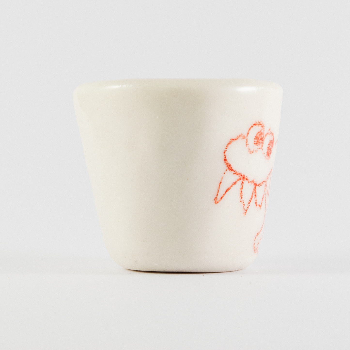 Side view of ceramic shot glass. There is red a hand-drawn doodle of a monster with large spikey teeth.