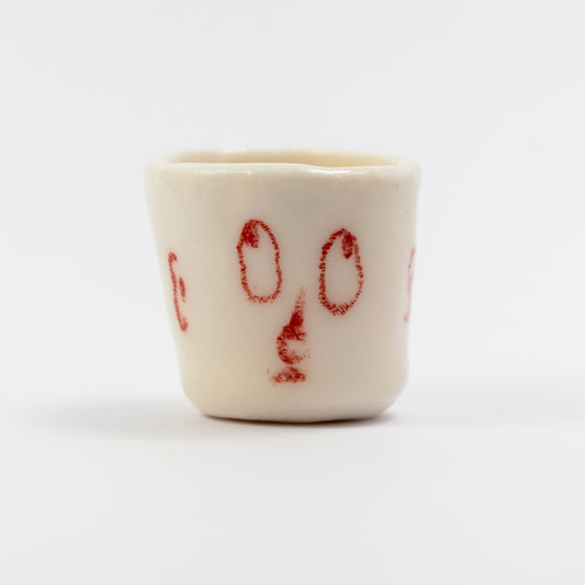 Small white porcelain cup with red underglaze pencil drawing of two oval eyes and a simple nose shape.