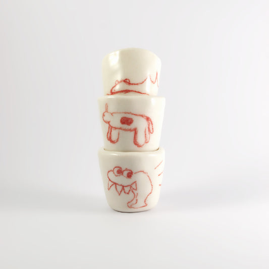 3 ceramic shot glasses stacked on top of each other. There is red hand-drawn doodles on each glass. Bottom: Monster with large spikey teeth. Middle: Horse like creature with circle on it's stomach. Top: A sleeping dinosaur.